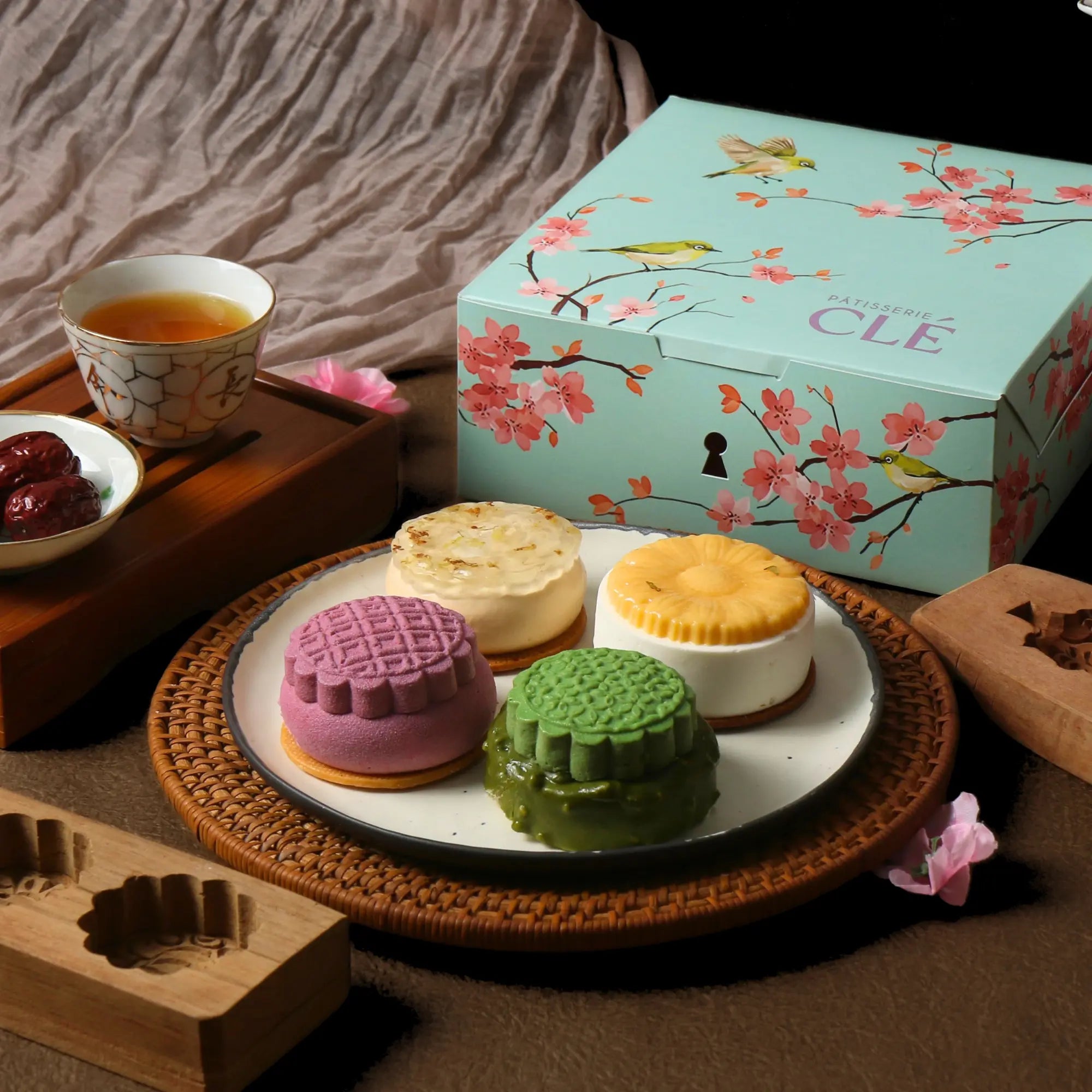 Four Pâtisserie CLÉ entremet mooncakes on a plate beside the signature mint cherry blossom gift box, styled with tea and jujubes.